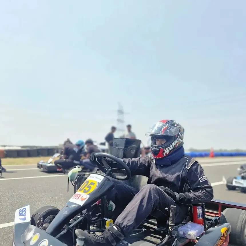 A driver in a full race suit and helmet beside the kart on track.